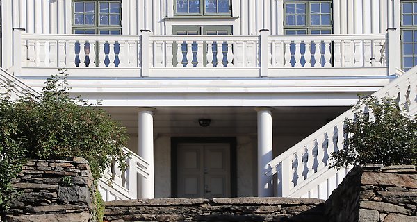 Detaljbild av vit herrgårdsbyggnad med balkong och trappor ned mot stenbelagd terrass.