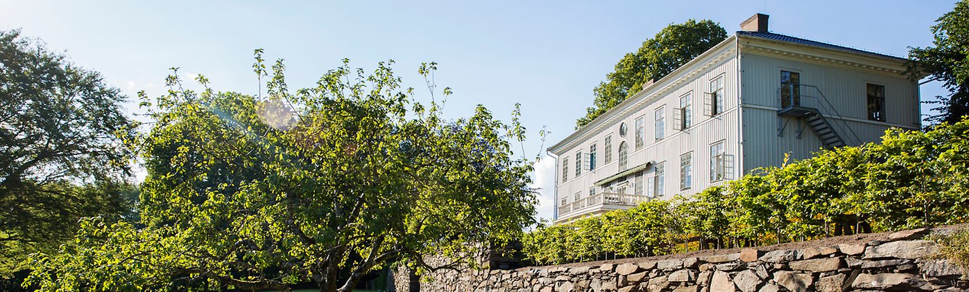 Den vita mangårdsbyggnaden på Råda Säteri fotograferad från sjösidan, med grönska och mur i förgrunden.